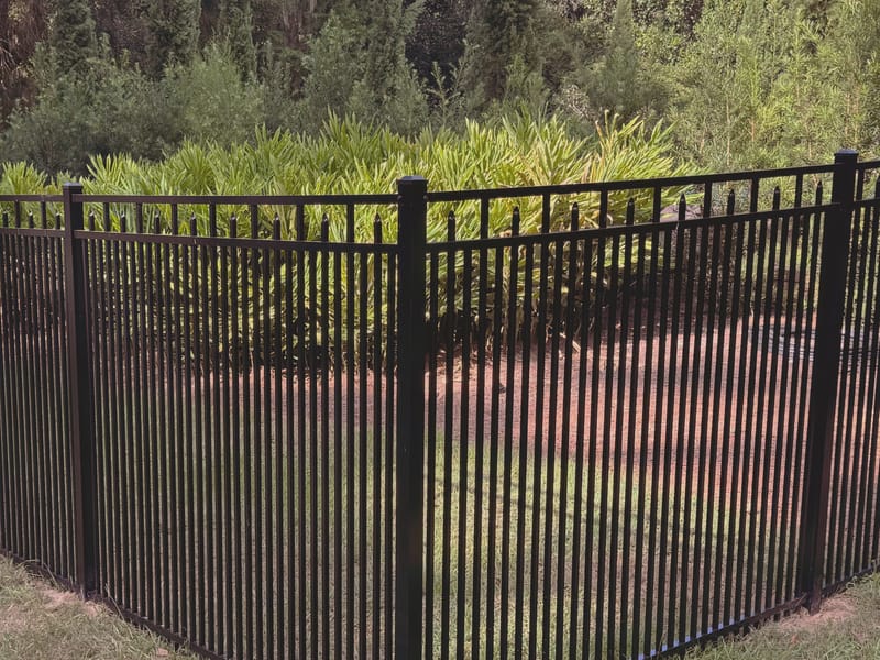 A black ornamental metal fence meeting at a right angle corner post, its narrow pickets rising past two flat rails to spear shaped tops, set on mown grass in front of dense green woodland.