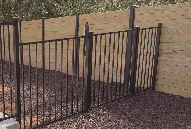 A black aluminum picket fence with a matching gate, set on a concrete edge in front of a horizontal board wood fence and a dark mulch bed.