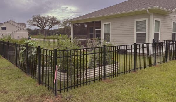 A black aluminum fence turning a corner behind a house, the flat top rail level across a slope.