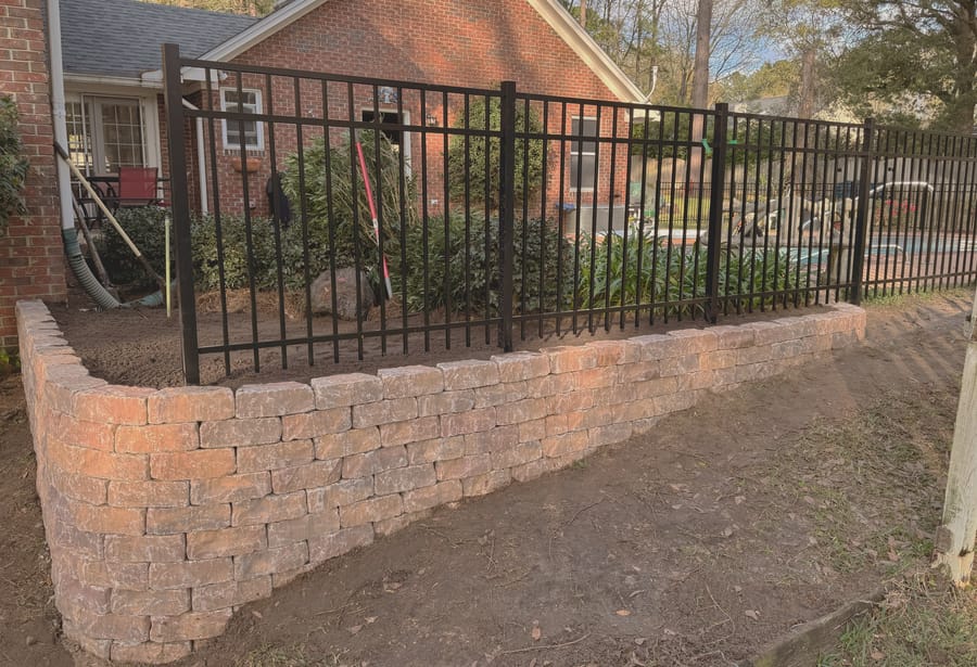 A black aluminum fence set along the top of a stacked stone retaining wall beside a brick house.