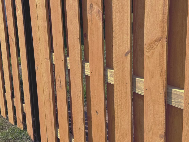 A black steel post set within a wood fence, with horizontal rails and vertical boards alternating front and back, receding along a lawn in raking sunlight.