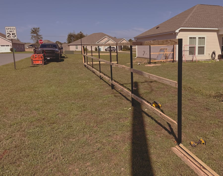 A fence part way through construction, steel posts up and the first rails attached, running along the side of a new house.