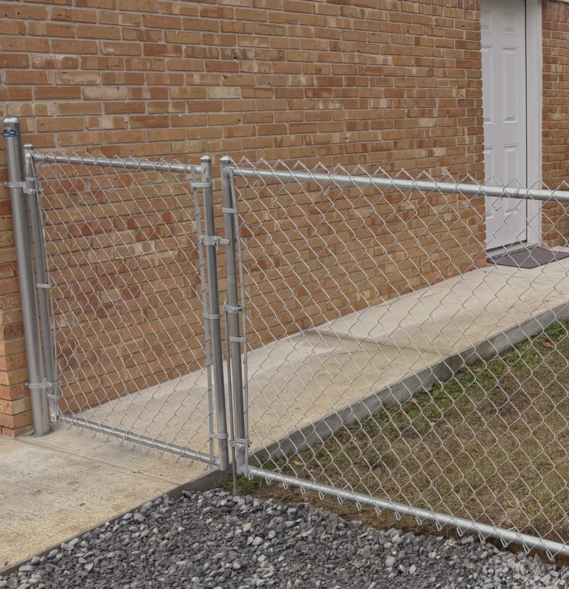 A galvanized chain link gate and fence beside a brick house, the mesh stretched between round posts with a concrete walkway behind it.