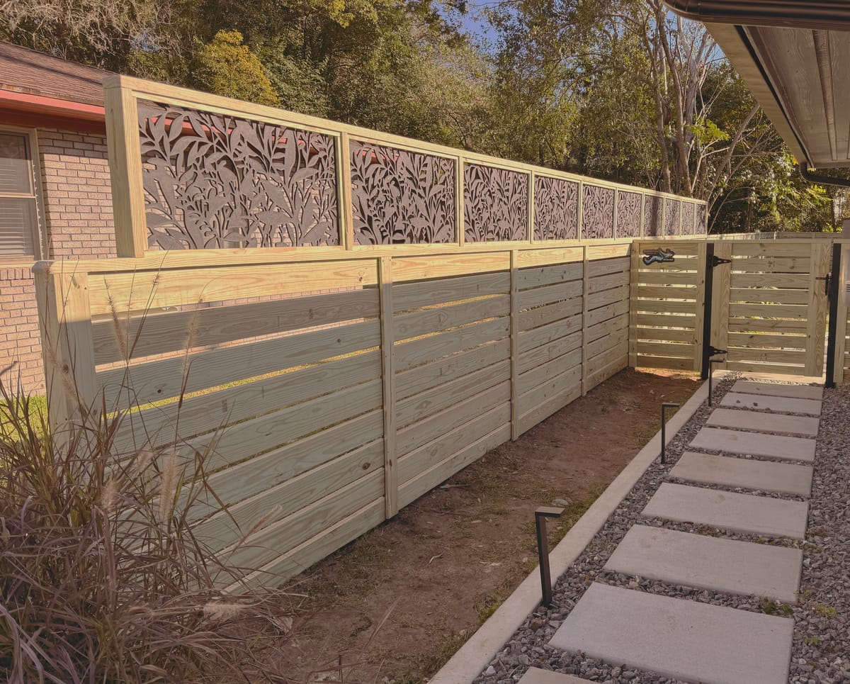 A horizontal slat fence with laser cut decorative metal panels set into the top section, running beside a stucco house past a stepping stone and gravel path.