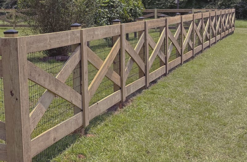 A wood fence of crossed diagonal braces set in square frames and backed with black wire mesh, turning at a capped corner post and running away across mown grass toward trees.