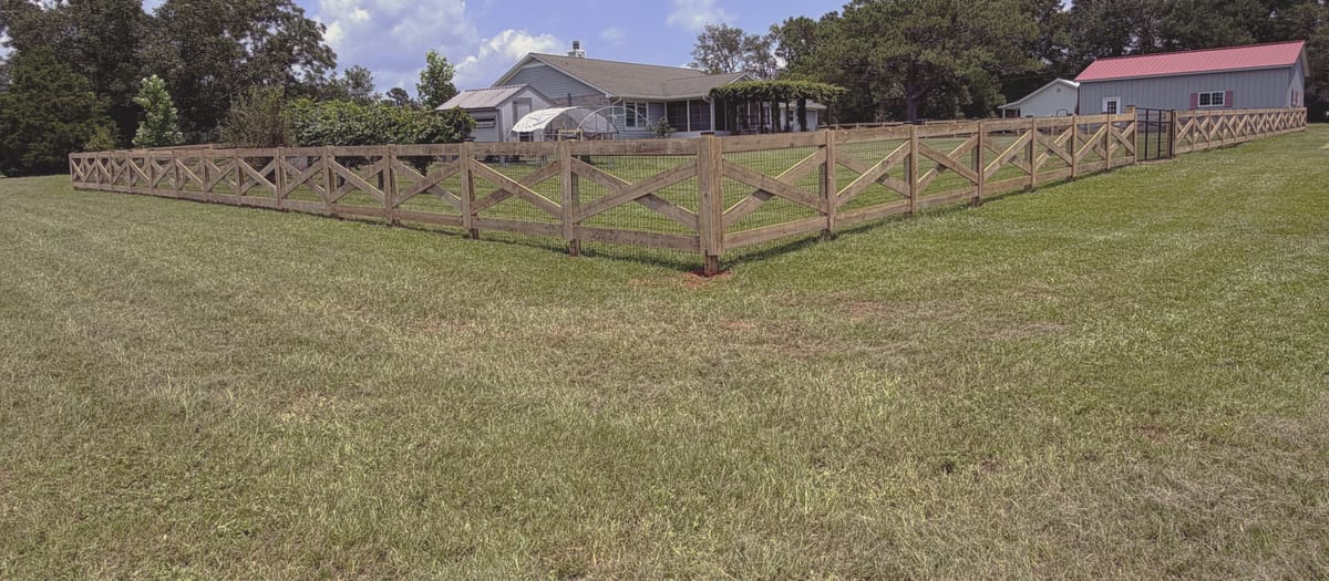 A crossbuck rail fence running the boundary of an open pasture with a house and outbuildings beyond.