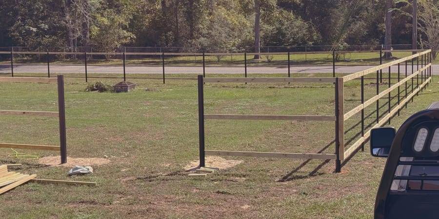 A black post and rail fence with wire infill enclosing a paddock, the line running away to a gate.