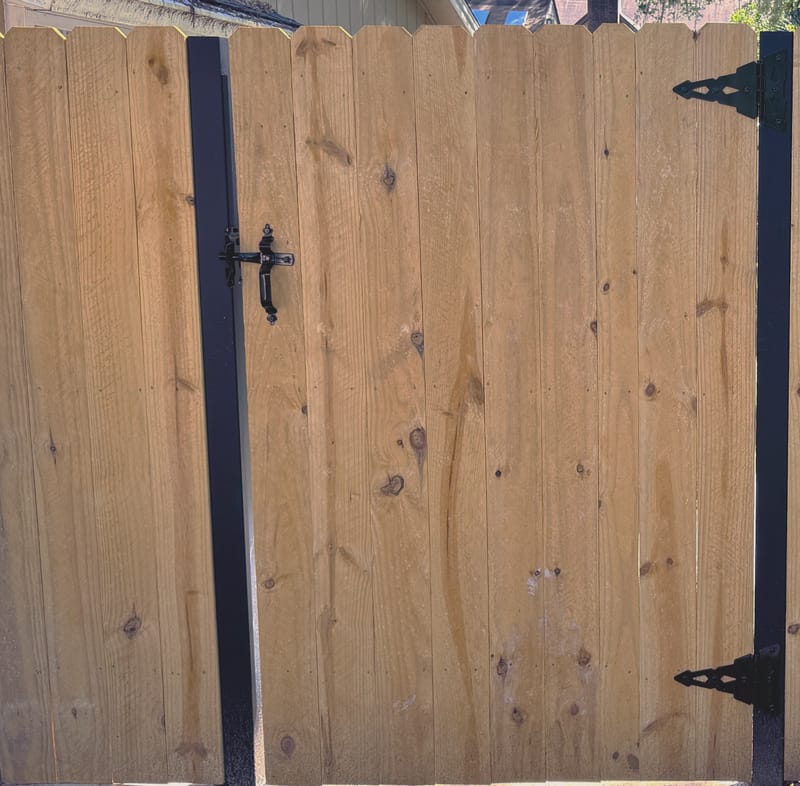 A wood gate with vertical boards and rounded tops, hung on black steel posts with black strap hinges and a black thumb latch handle, set over a concrete pad.