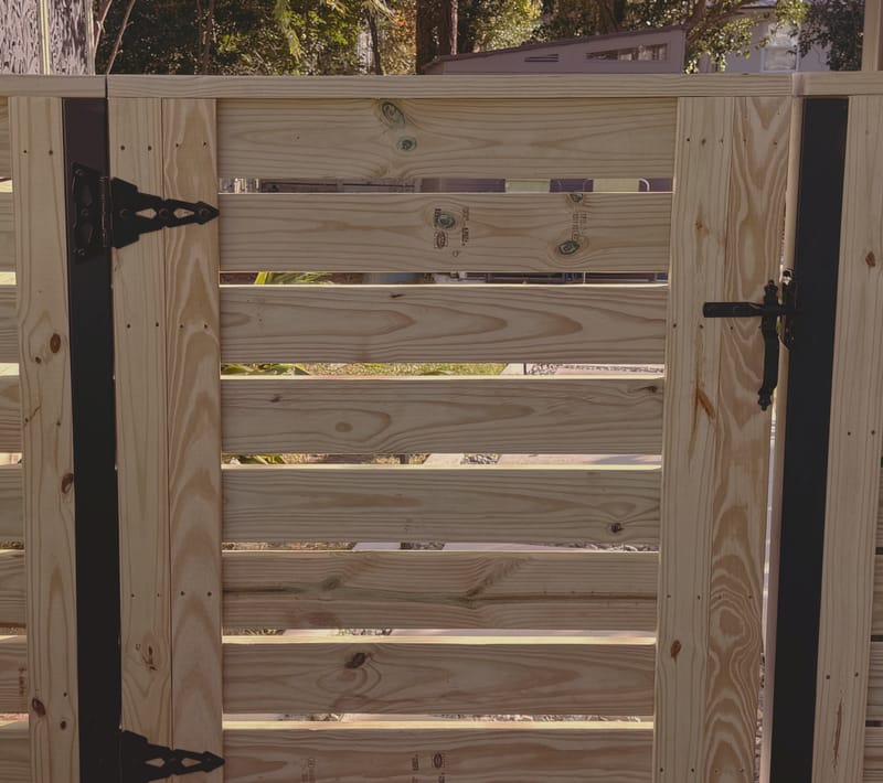 Two horizontal slat wood gate leaves on black steel posts, with ornate black strap hinges on the centre post and a black latch with a drop rod on the adjoining leaf.