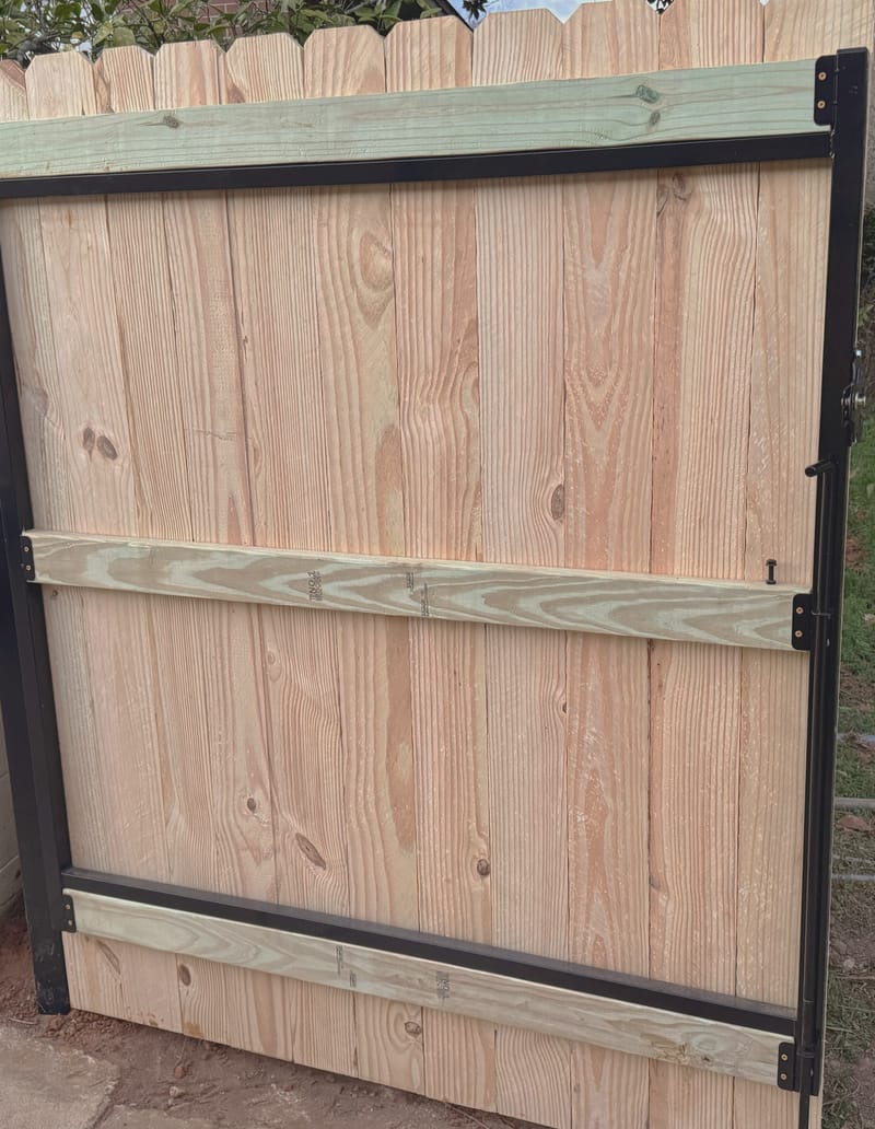 A gate leaf part way through assembly: a black steel perimeter frame with timber rails across the top, middle and bottom, filled with vertical boards, and black hinges and a latch fitted to one stile.