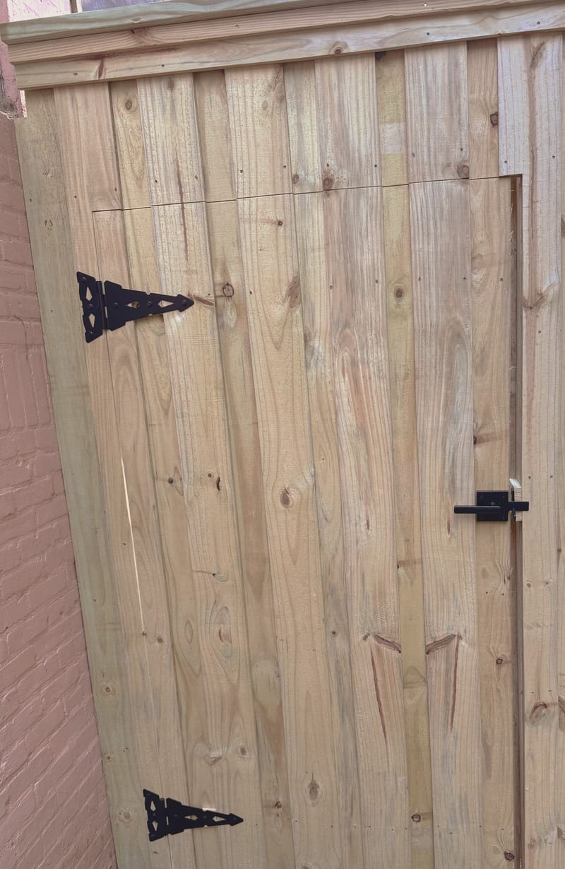 Close view of a wood gate built from vertical boards, with two black strap hinges and a black lever latch, framed by a capped top rail and set against a painted brick wall.