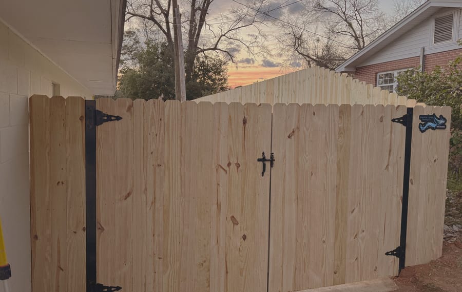 A wood double gate across a driveway opening, with black strap hinges and a drop rod at the center.