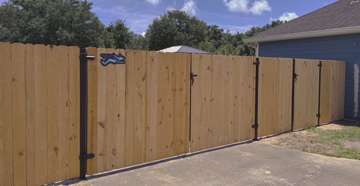 A wood double drive gate closed across a concrete driveway, hung on black steel posts with heavy strap hinges.