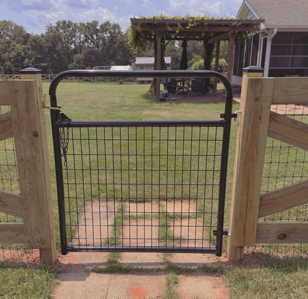 A welded wire garden gate in a black steel frame between two heavy wood posts, opening on to a lawn and a pergola.