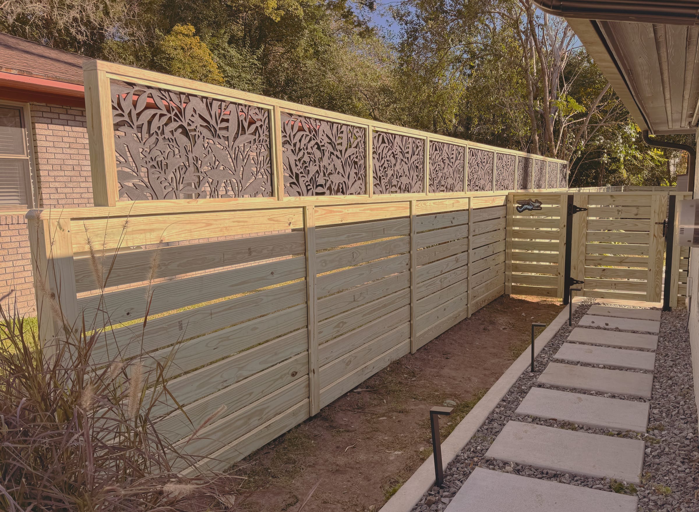 A horizontal slat fence with laser cut metal panels set into the upper section, running beside a house past a stepping stone path.
