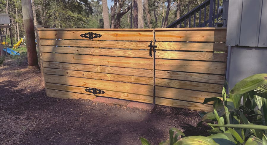 A low horizontal board fence with black strap hinges and a drop latch, set among oaks.