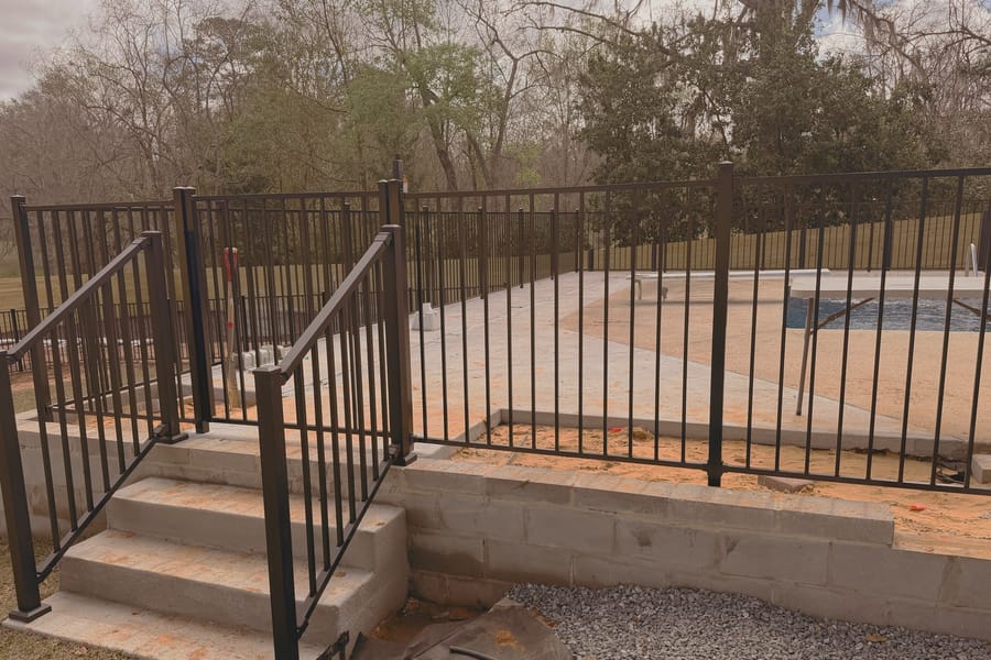 A black aluminum pool fence and matching handrail beside pool steps, with the pool deck under construction.