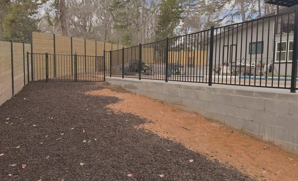 A black aluminum fence mounted along the top of a block retaining wall above a pool deck.