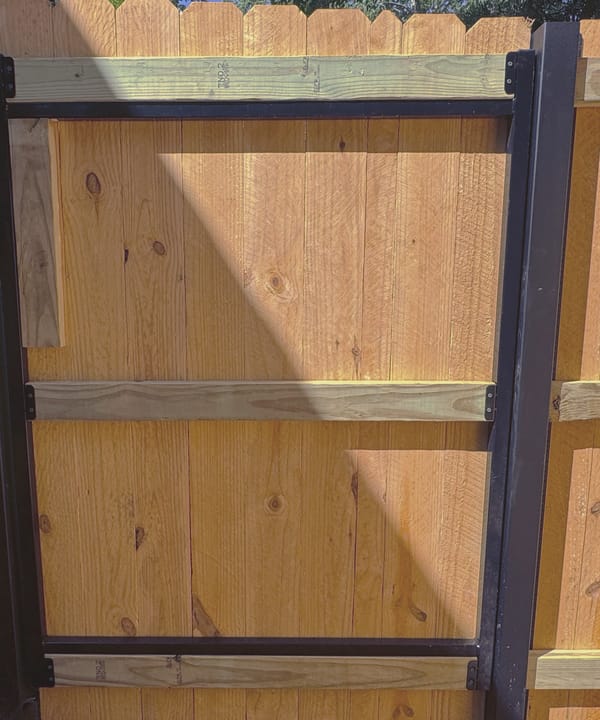 Close view of a black steel fence post with a pressure treated rail bolted to it and a welded steel gate frame beside it.