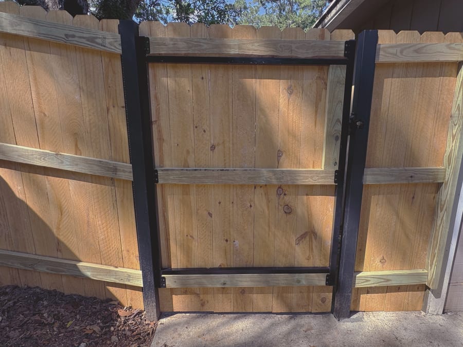 The back of a wood privacy fence, showing black steel posts with the horizontal rails bolted through to them and a welded steel gate frame hung on the same posts.