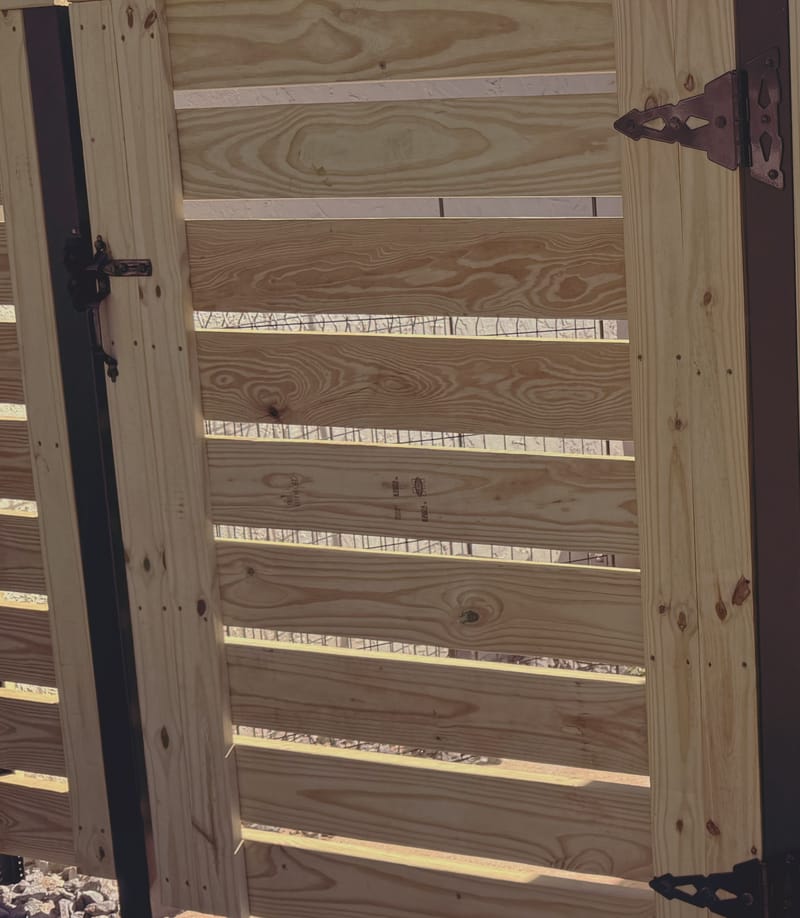 A gate built from horizontal boards with narrow gaps between them, hung on black steel posts with heavy black strap hinges and a black pull handle, continuing into a matching fence.
