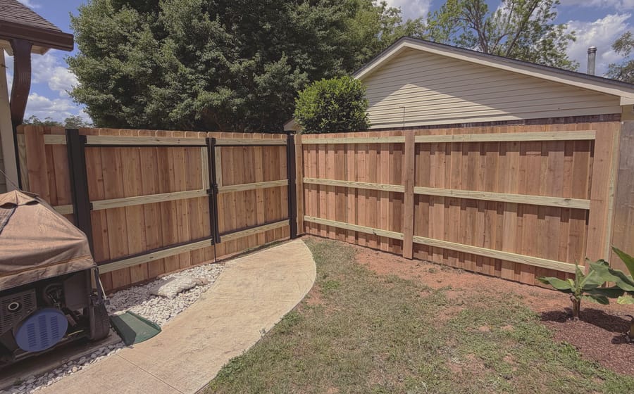 A new wood fence meeting an older weathered run at a gate, with a curved concrete walk between them.